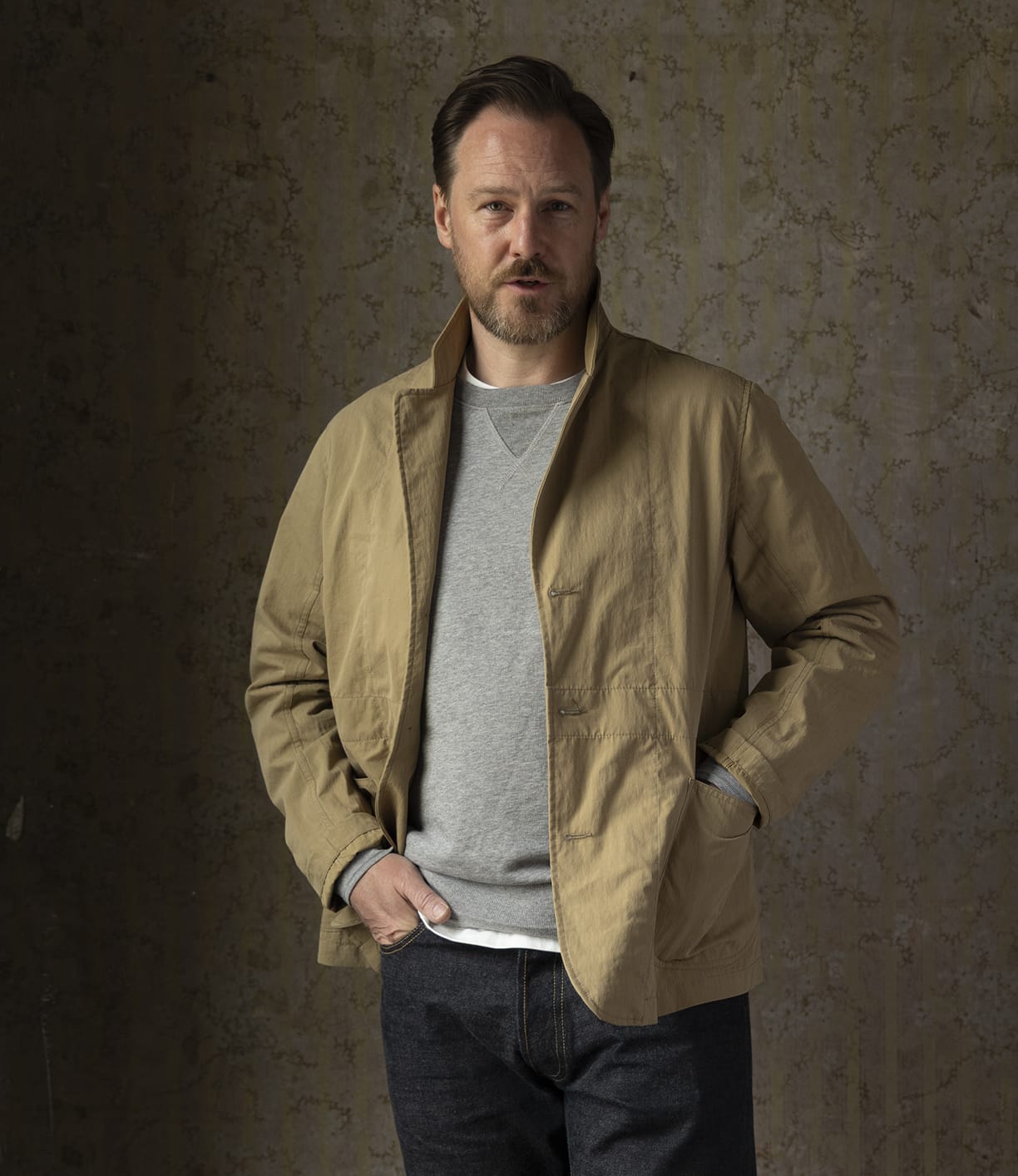 A man with short brown hair and a beard stands before a textured brown wall, wearing the Best Made Co Possibilities Jacket in Tan over a gray sweatshirt and organic cotton selvedge denim jeans, one hand in his pocket, the other at his waist.