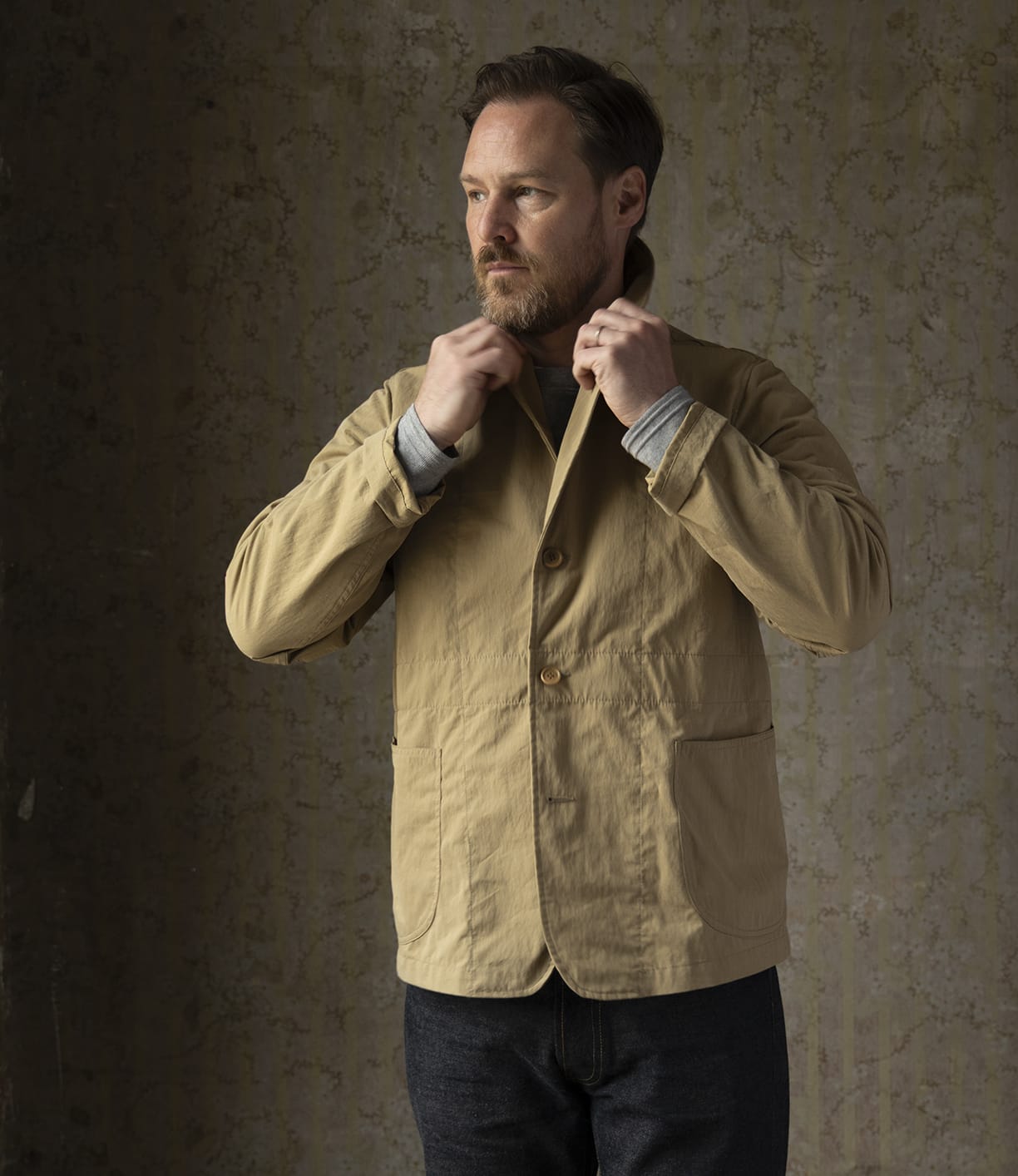 A bearded man adjusts the collar of his Possibilities Jacket Tan by Best Made Co while looking to the side. He stands against a textured, neutral background, wearing a gray shirt and dark pants made from organic cotton selvedge denim.