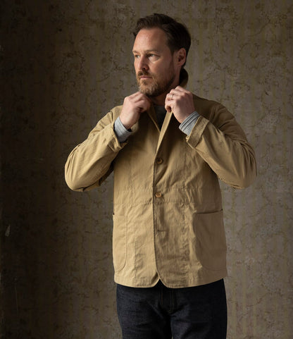 A man with a beard and short hair stands indoors against a textured wall, adjusting the collar of his Best Made Co. Possibilities Jacket Tan layered over a gray shirt and dark pants, looking off to the side.