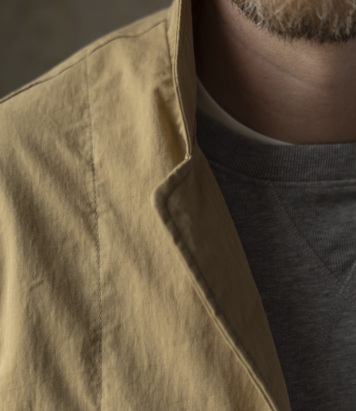 Close-up of a person's neck and upper chest wearing the Best Made Co Possibilities Jacket Tan, layered over a gray crew-neck sweatshirt. The person's short facial hair is partially visible.