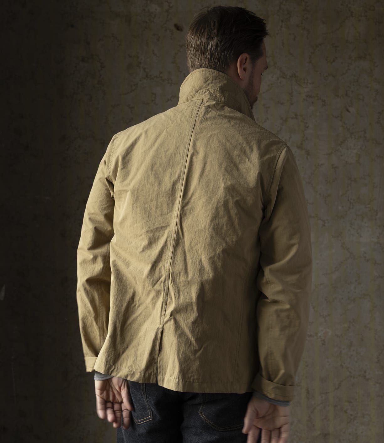 A man stands with his back to the camera, wearing the Best Made Co Possibilities Jacket Tan with the collar up and dark organic cotton selvedge denim jeans, posed against a textured neutral wall—showcasing American craftsmanship.