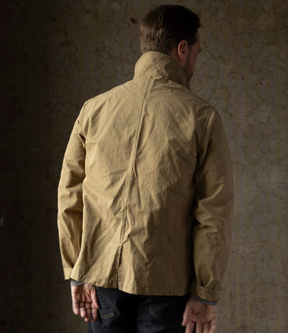 A person stands facing a textured wall, wearing Best Made Co.’s Possibilities Jacket Tan with the collar turned up and dark jeans. Their hands are visible behind their back.