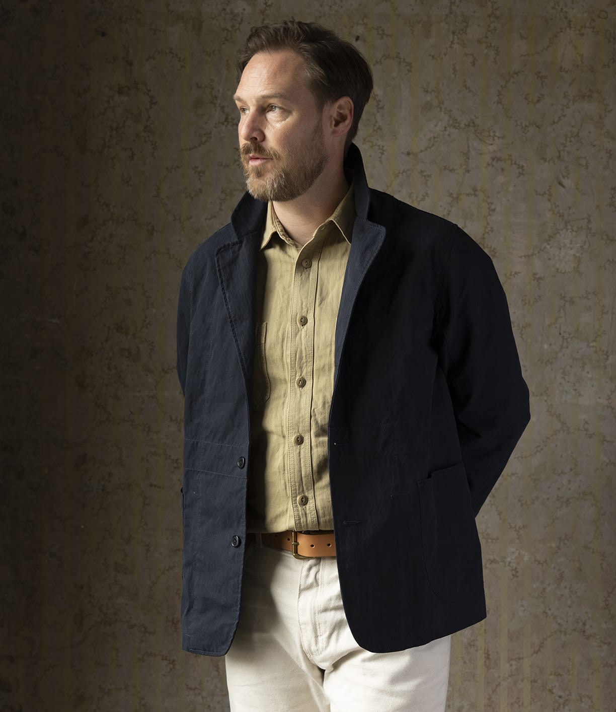 A man with light brown hair and a beard, wearing the Best Made Co Possibilities Jacket Navy over a beige shirt and light trousers with a tan belt, stands against a textured brown background with his hands behind his back, looking left.