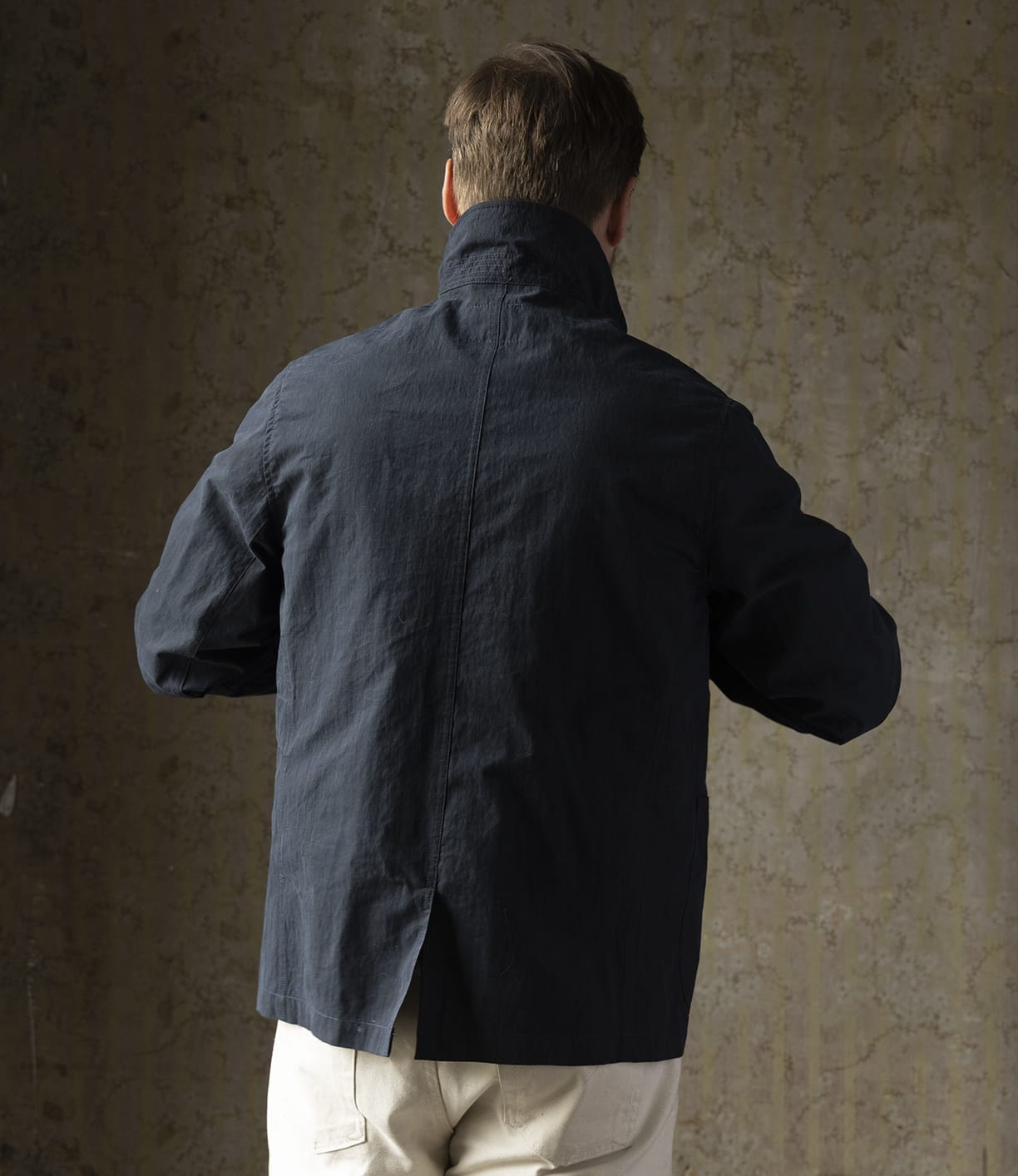 A person with short brown hair is shown from behind, wearing a Best Made Co Possibilities Jacket Navy and off-white pants, standing against a textured, neutral-colored wall.