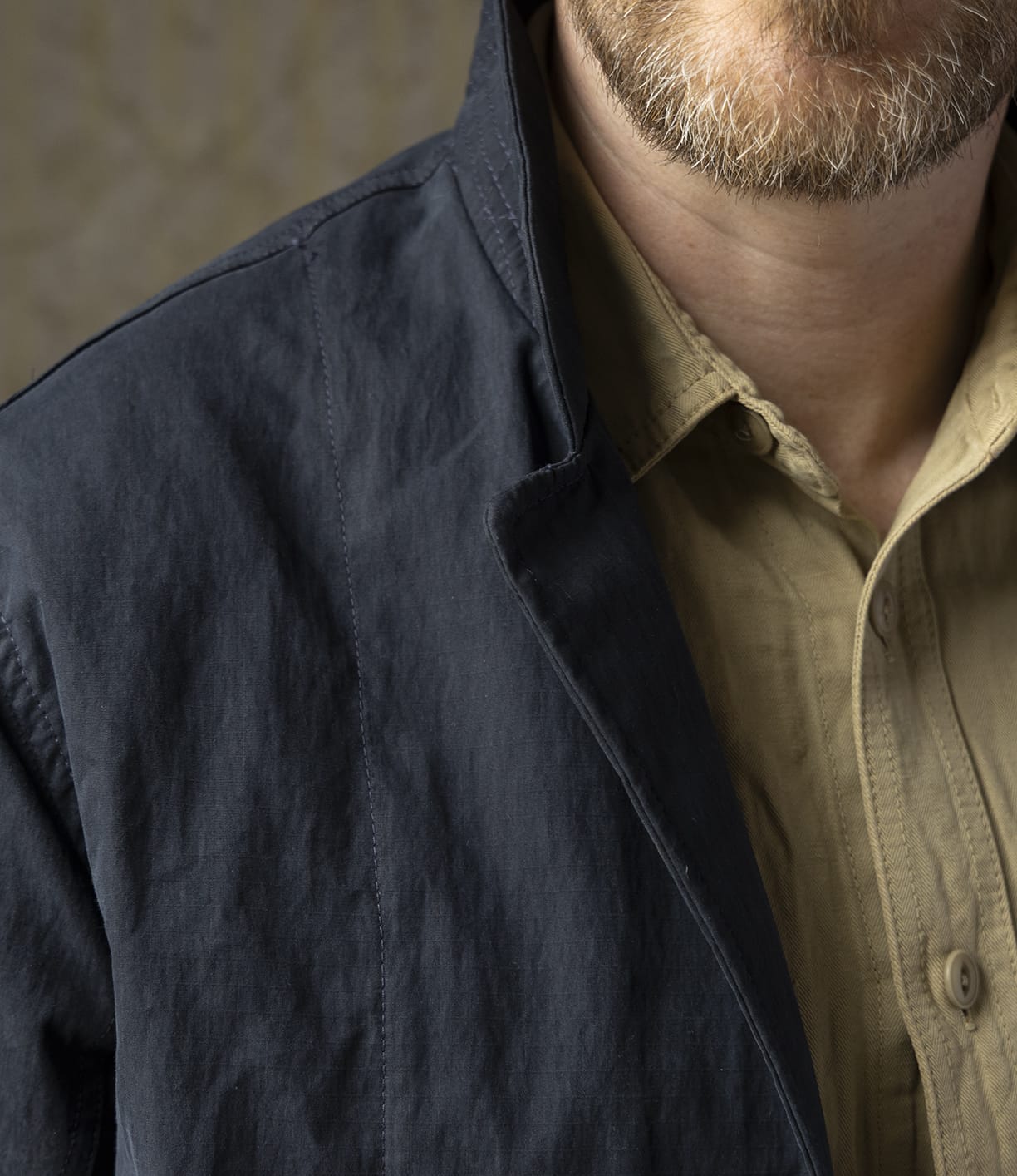 A close-up shows a person with a light brown beard wearing a beige button-up shirt and the navy Possibilities Jacket by Best Made Co, highlighting American craftsmanship. Only the lower half of the face, neck, and upper torso are visible.