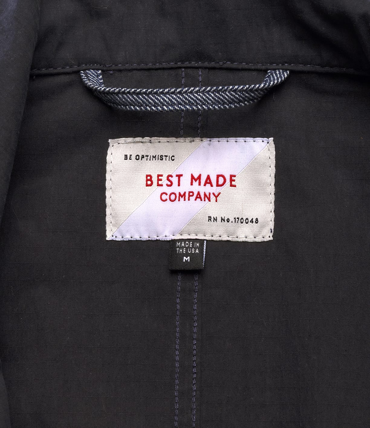 A close-up of the Possibilities Jacket Navy by Best Made Co shows the label reading: BEST MADE COMPANY, BE OPTIMISTIC, Made in the USA, size M, with detailed stitching that highlights American craftsmanship.