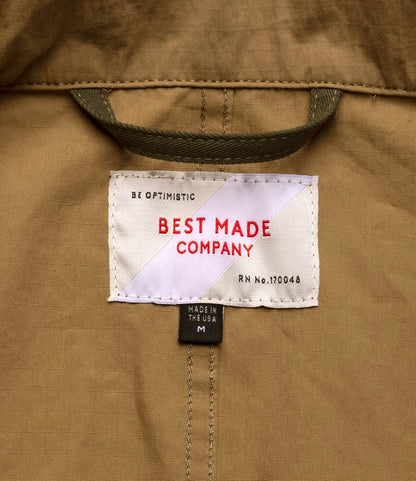 A close-up of a label on tan fabric from the Best Made Co. Possibilities Jacket Tan, reading BEST MADE COMPANY in red, BE OPTIMISTIC above, and smaller tags below that state MADE IN THE USA and M.