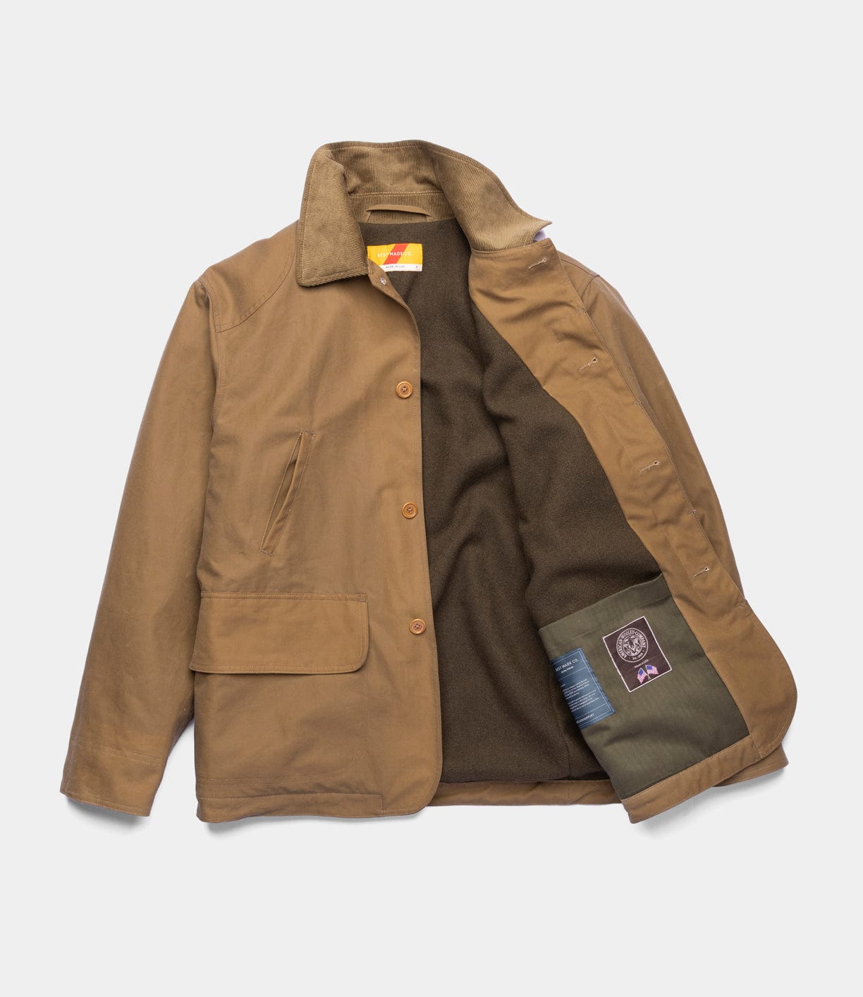 The Best Made Co. Wool-Lined Waxed Field Jacket in tan is weather-resistant, featuring a corduroy collar, button front, large front pocket, dark brown fleece lining, and an inner green pocket with a purple label.