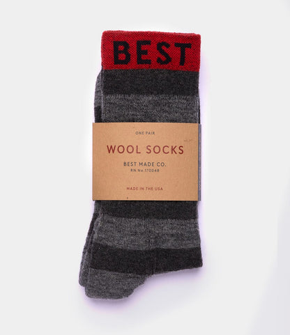 The Standard Wool Socks by Best Made Co. feature gray and dark gray stripes, a red BEST band at the top, and come wrapped in a brown “WOOL SOCKS, BEST MADE CO., MADE IN THE USA” label. Enjoy all-season comfort with these stylish merino wool socks.