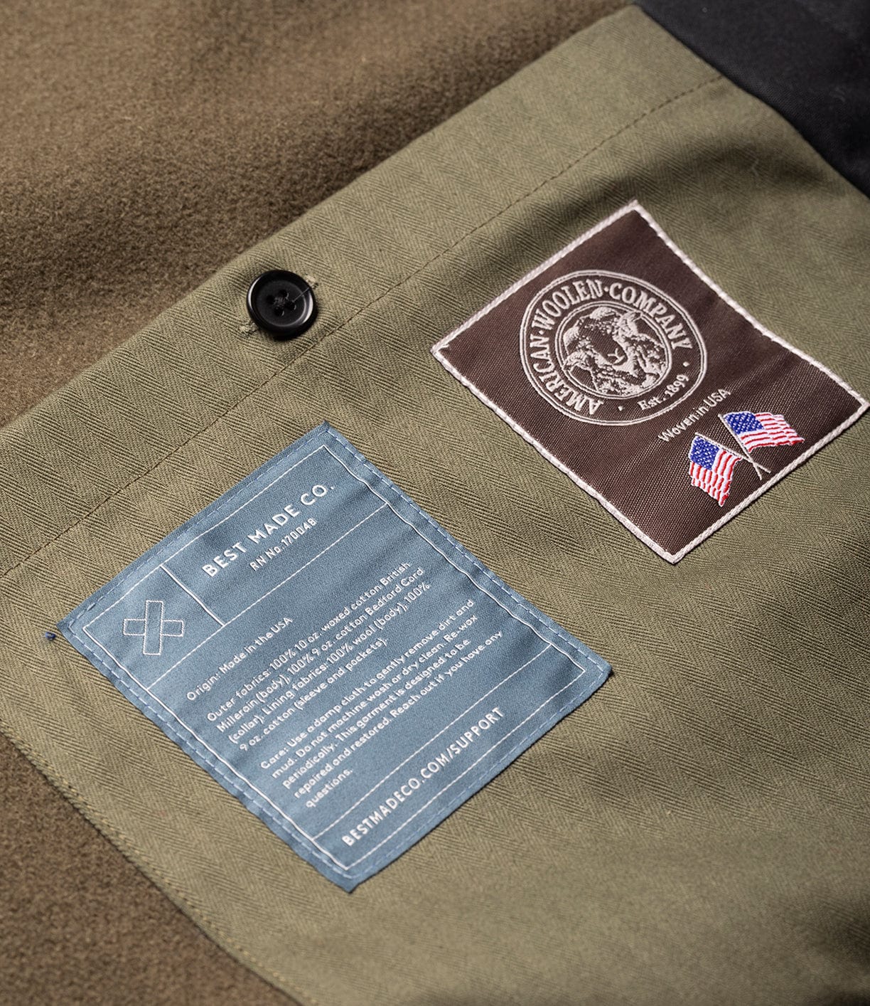 Close-up of black fabric on the Wool-Lined Waxed Field Jacket by Best Made Co., showing two sewn labels (one from the American Woolen Company with U.S. flags, one from Best Made Co. with product info) and a black button.