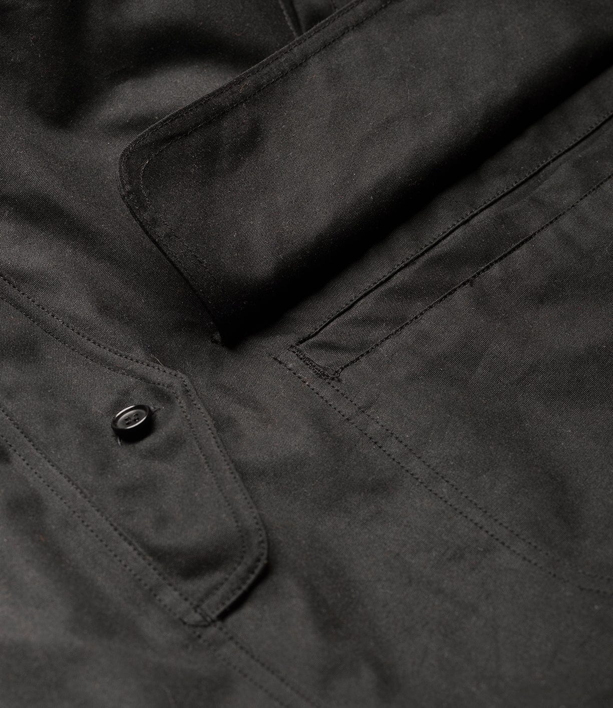 Close-up of the Best Made Co. Wool-Lined Waxed Field Jacket Black, showing a flap pocket with stitched outline and a single black button. The British Millerain Staywax material's texture and detailed stitching are clearly visible.