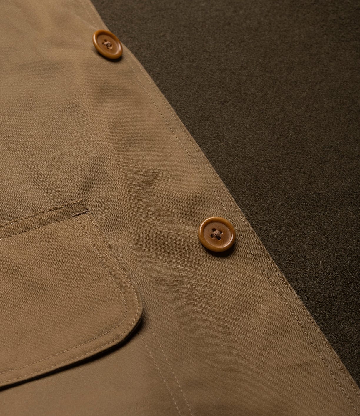 Close-up of the Best Made Co. Wool-Lined Waxed Field Jacket in Tan, featuring a smooth, slightly reflective weather-resistant fabric, a pocket, and two brown buttons, laid on a dark olive green background.