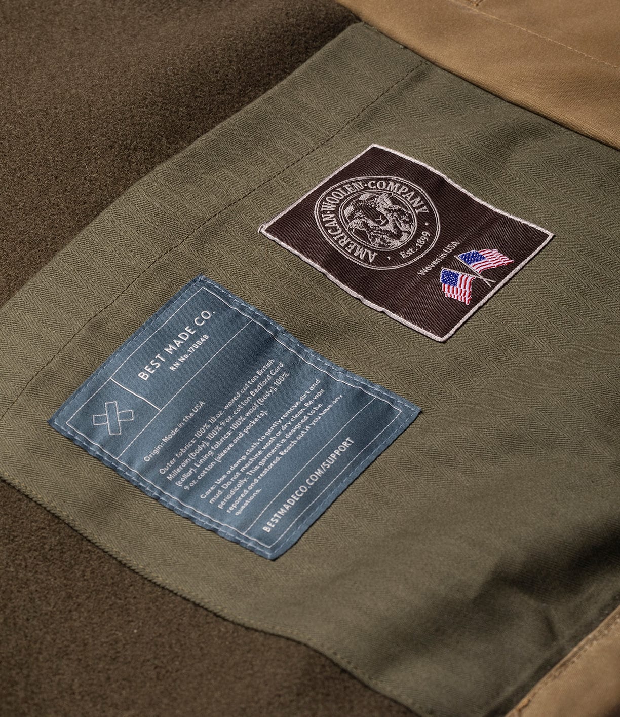 Close-up of the Wool-Lined Waxed Field Jacket Tan by Best Made Co., showing the inside with two sewn-on labels: one black with a circular logo and American flags, and another blue label with white care instructions.