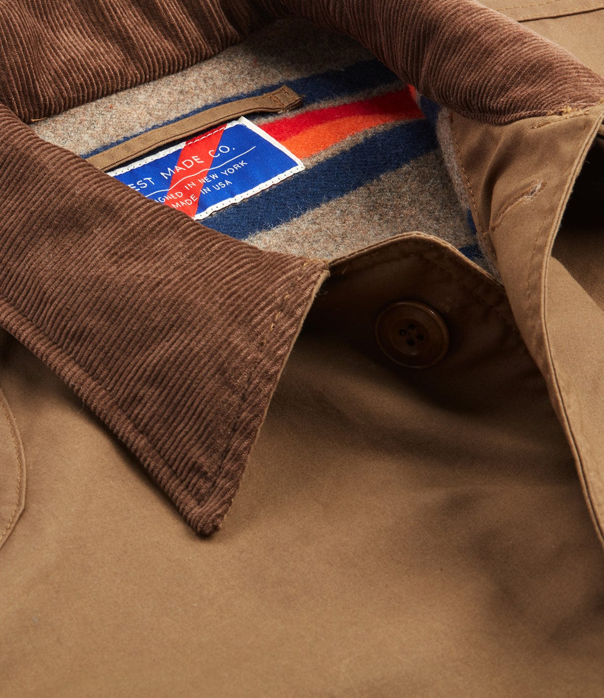 Close-up of the Best Made Co. Wool-lined Waxed Field Jacket in Tan: features a brown waxed finish, corduroy collar, colorful striped wool lining, button closure, and a blue-and-red interior label—classic all-weather outerwear.