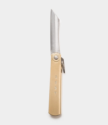 The Mini Brass Higo Knife by Best Made Co. features a metallic blade and a beige handle engraved with "BEST," inspired by Japanese folding knife tradition and handmade Edo-period design, shown on a plain white background.