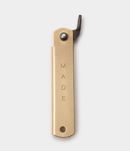 The Best Made Co. Mid Brass Higo Knife features "MADE" engraved vertically on its brass handle, a curved black metal opener at the top, and a bottom hole, reminiscent of traditional Japanese folding knives, set on a plain light background.