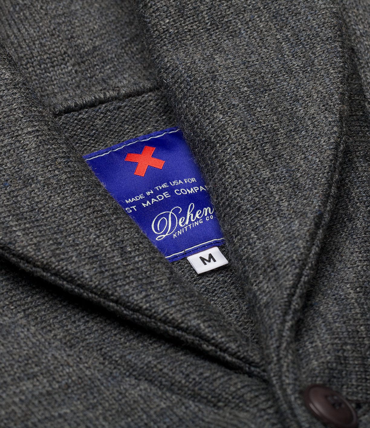 Close-up of the Dehen Shawl Neck Sweater Charcoal by Best Made Co. A blue label inside the gray textured pocket reads "Made in the USA, Dehen Knitting Co., Portland Oregon sweater," with a red X and M size tag.