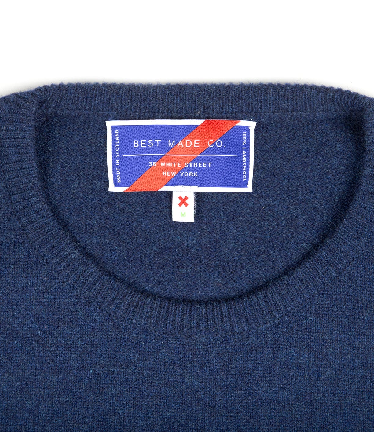 A close-up of the Lightweight Shetland Sweater's blue crew neck collar reveals a "Best Made Co. 36 White Street, New York" label with a red stripe, plus a smaller M size tag with a red X, crafted in partnership with Scottish woolen mills.